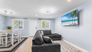 A living room with a sectional sofa, a dining table with chairs, a TV mounted on the wall, and a patterned rug.