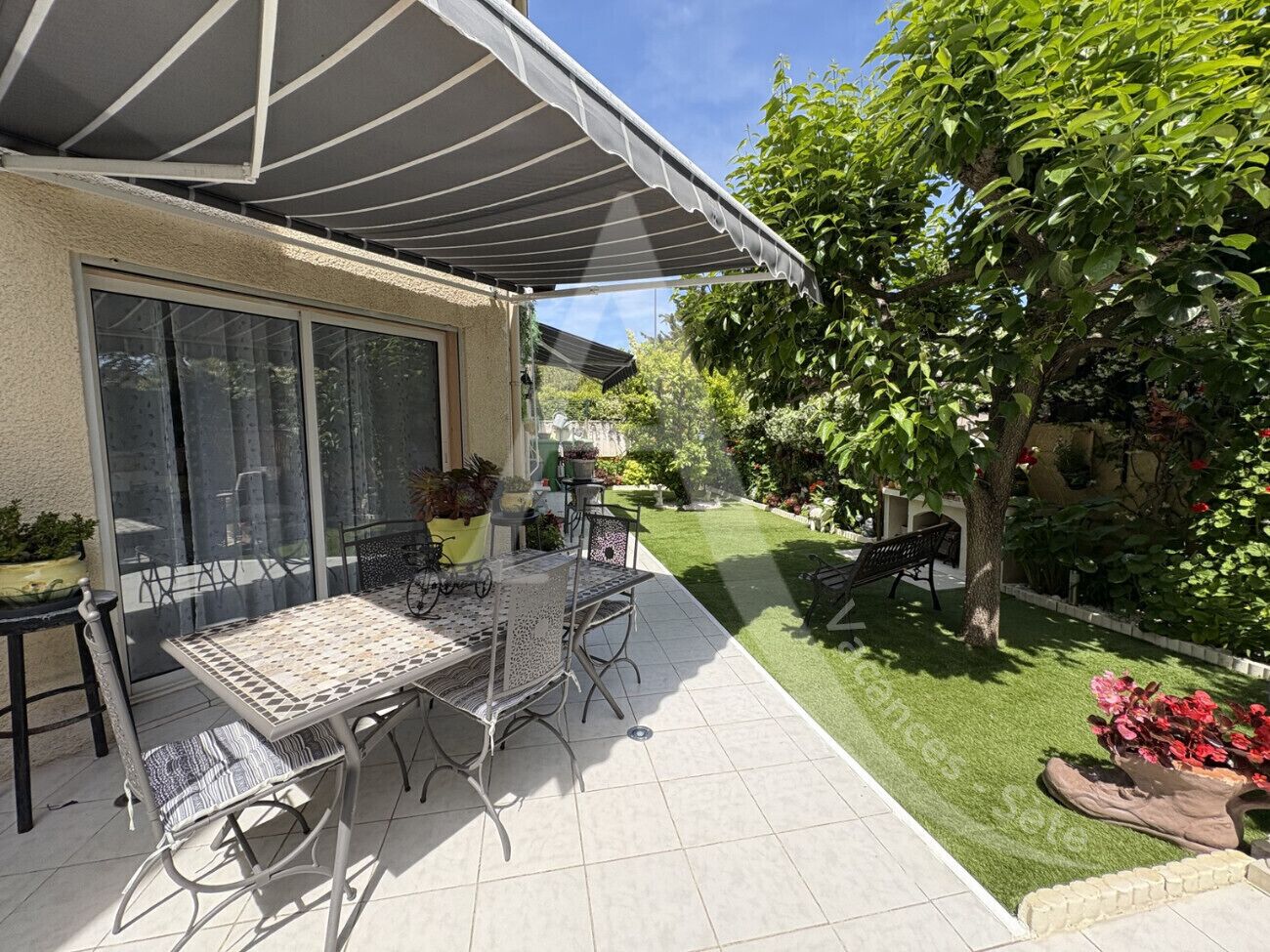 A patio with a table and chairs, a covered area, and a garden with a lawn and trees.