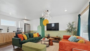 A modern living room with a green sofa, an orange sofa, and a green ottoman. There is a flat-screen TV mounted on the wall, a coffee table, and a potted plant. - Heart of Dallas in Knox Henderson | Sleeps 4 (Dallas)