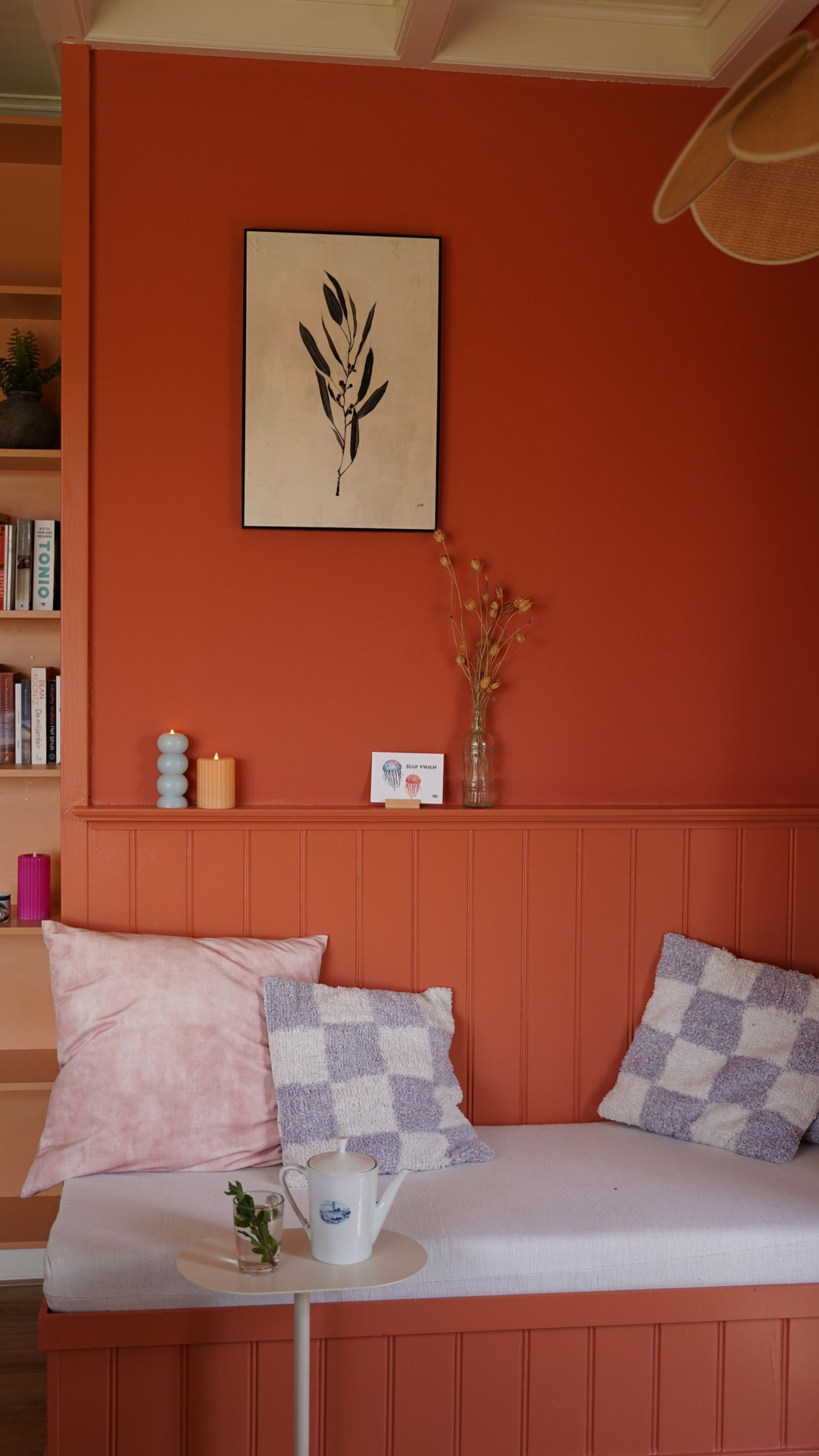 Ein Zimmer mit einer Couch, Kissen, einem kleinen Tisch mit einer Tasse, einer Vase mit Trockenblumen, einem gerahmten Kunstwerk und einem Bücherregal.