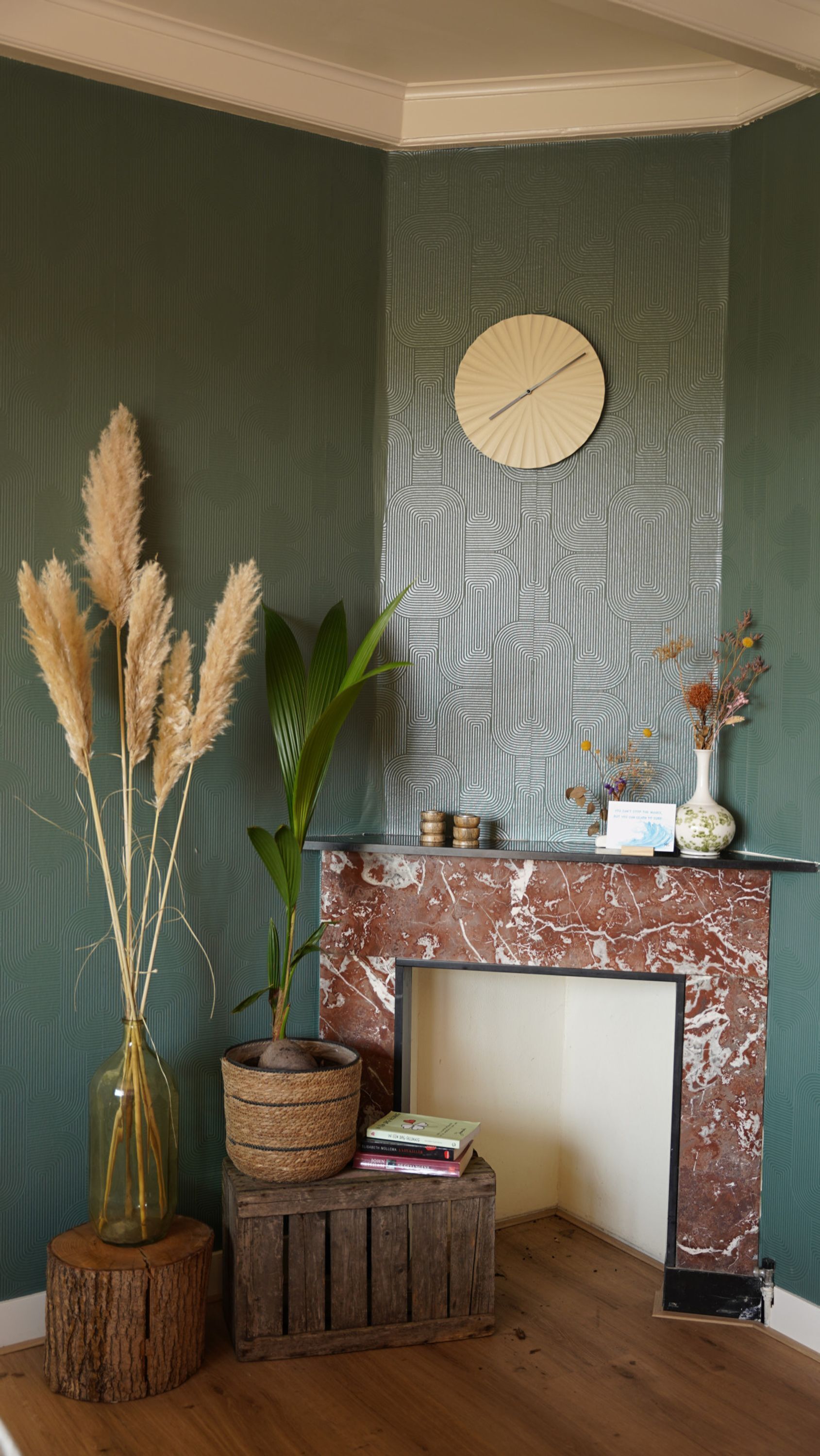 A corner room with a green patterned wall, a wooden mantel with books and decorative items, a round wall clock, and a rustic wooden stool.