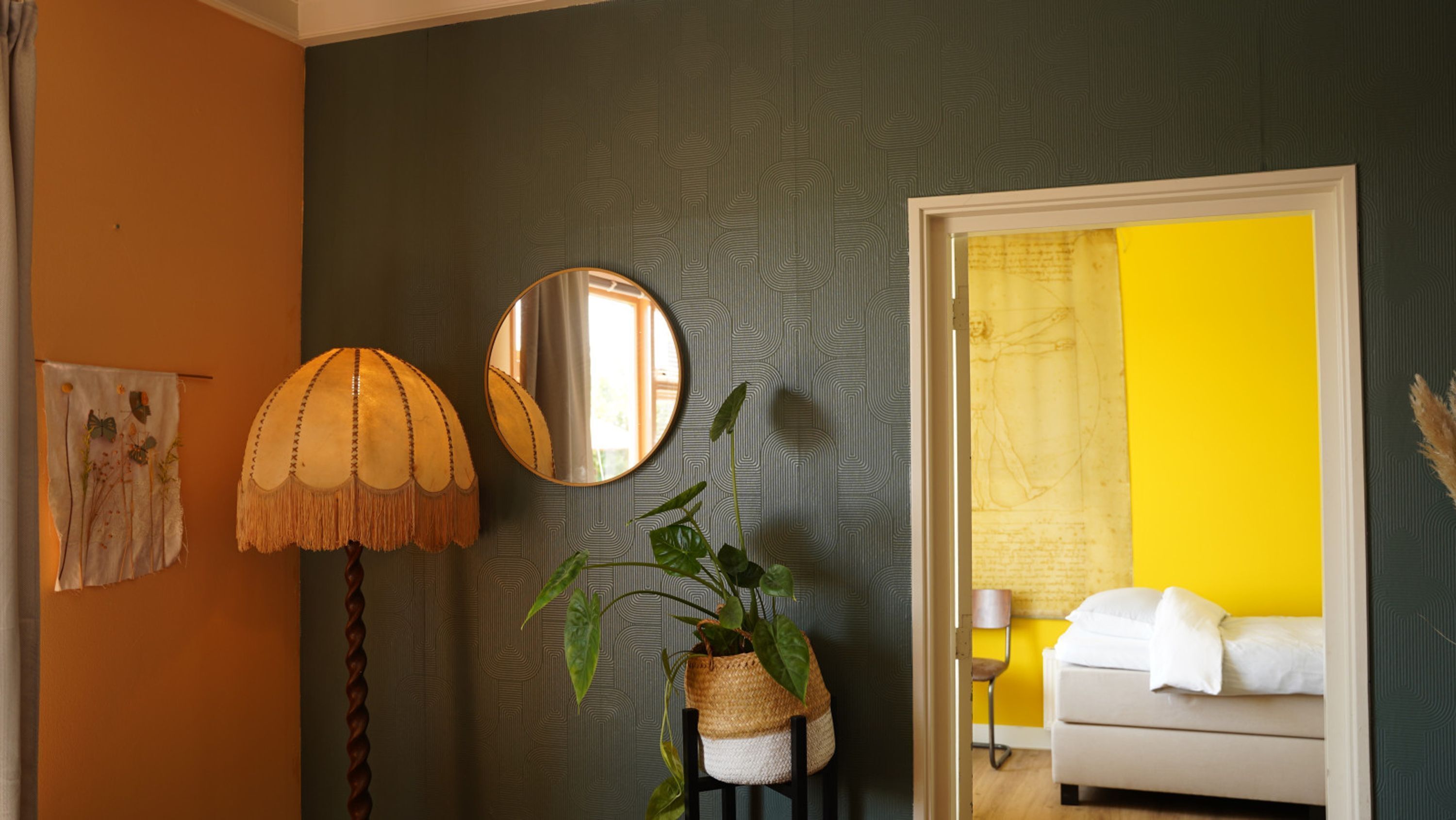 Ein Zimmer mit Wandmuster, einem runden Spiegel, einer Pendelleuchte, einer Topfpflanze und einem Blick auf ein Bett vor einer gelben Wand im Hintergrund.