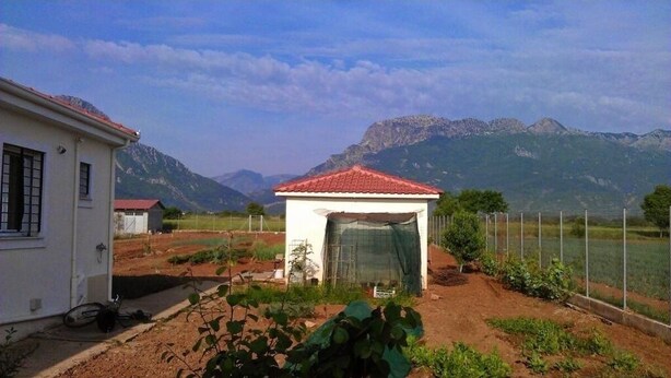 Une maison de campagne avec un toit recouvert de tuiles rouges, un petit jardin et des montagnes en arrière-plan.