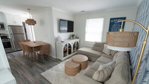 Una sala moderna con un sofá beige, una mesa de comedor con sillas de mimbre, una cocina con refrigerador de acero inoxidable y un televisor de pantalla plana montado en la pared.