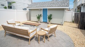 Un patio con un banco de madera, dos sillas y una pequeña mesa. Se ven dos plantas en macetas y una puerta azul al fondo.