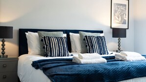 A neatly made bed with blue and white striped pillows, a dark blue headboard, and a white wall with a framed picture. - Woodside Family Pet-friendly Holiday Home With Hot Tub (Fraserburgh)