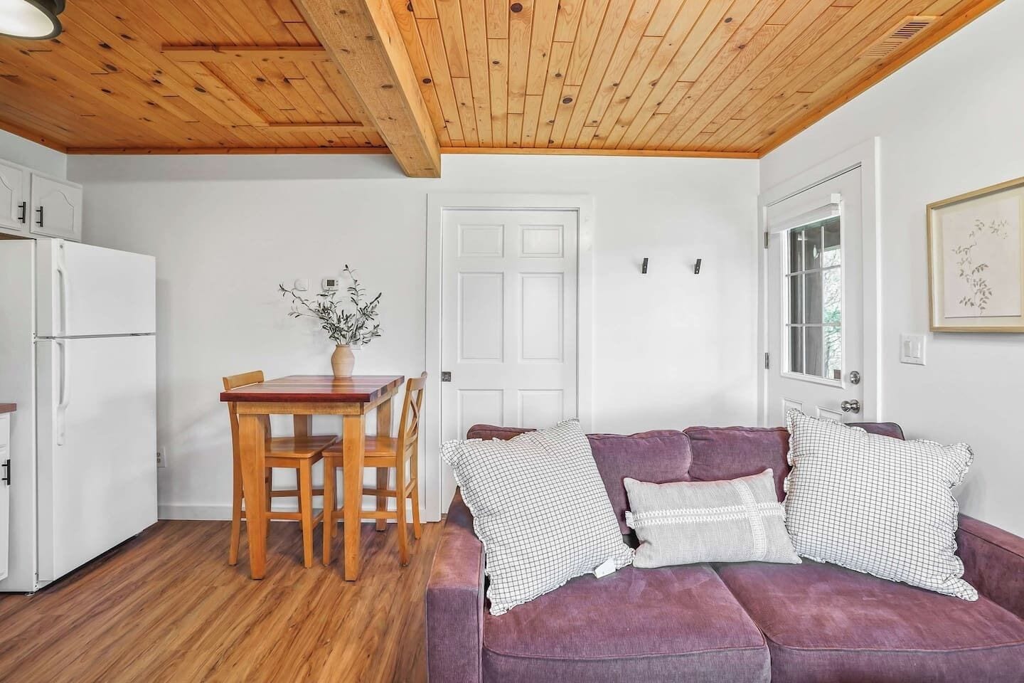 A compact living space with a wooden ceiling, a purple sofa with patterned cushions, a wooden dining table with chairs, and a white door.