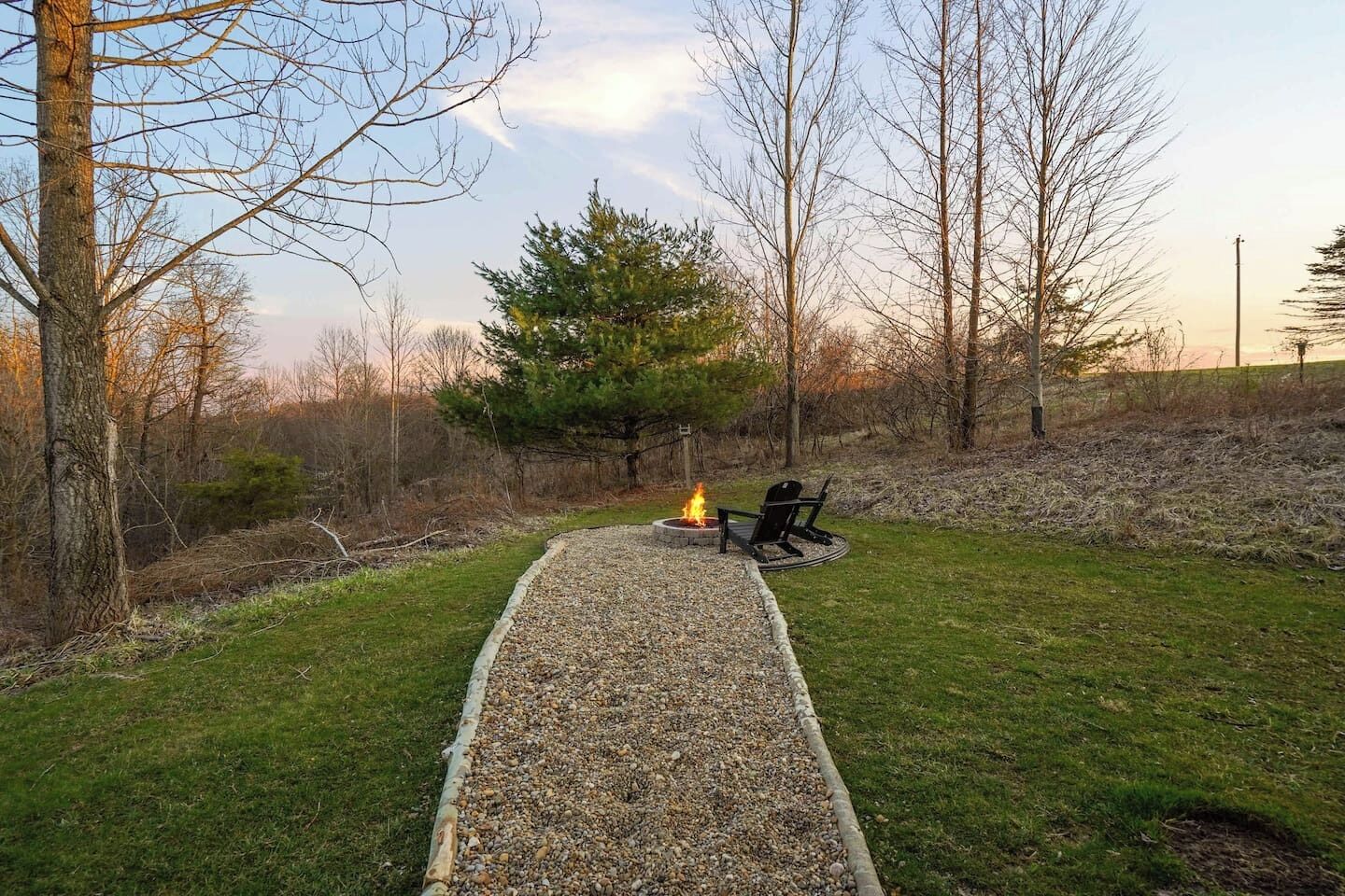 A gravel path leading to a fire pit with two chairs, surrounded by trees and grass.
