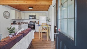 A compact kitchen with white cabinets, a microwave, and an oven. There is a wooden dining table with two chairs. A clock is mounted on the wall, and a plant is on the floor.