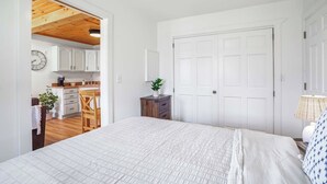 A bedroom with a large bed, a wooden ceiling, a white door, and a kitchen visible through an open doorway.