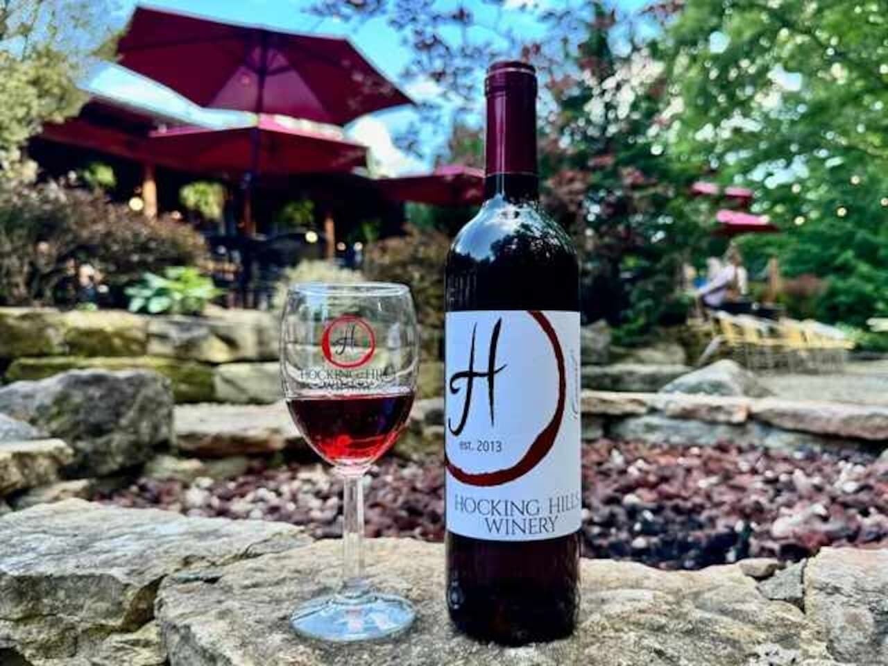 Una bottiglia di vino e un bicchiere su una superficie in pietra, con un'etichetta che recita “Hocking Hills Winery” e “fondata nel 2013”.