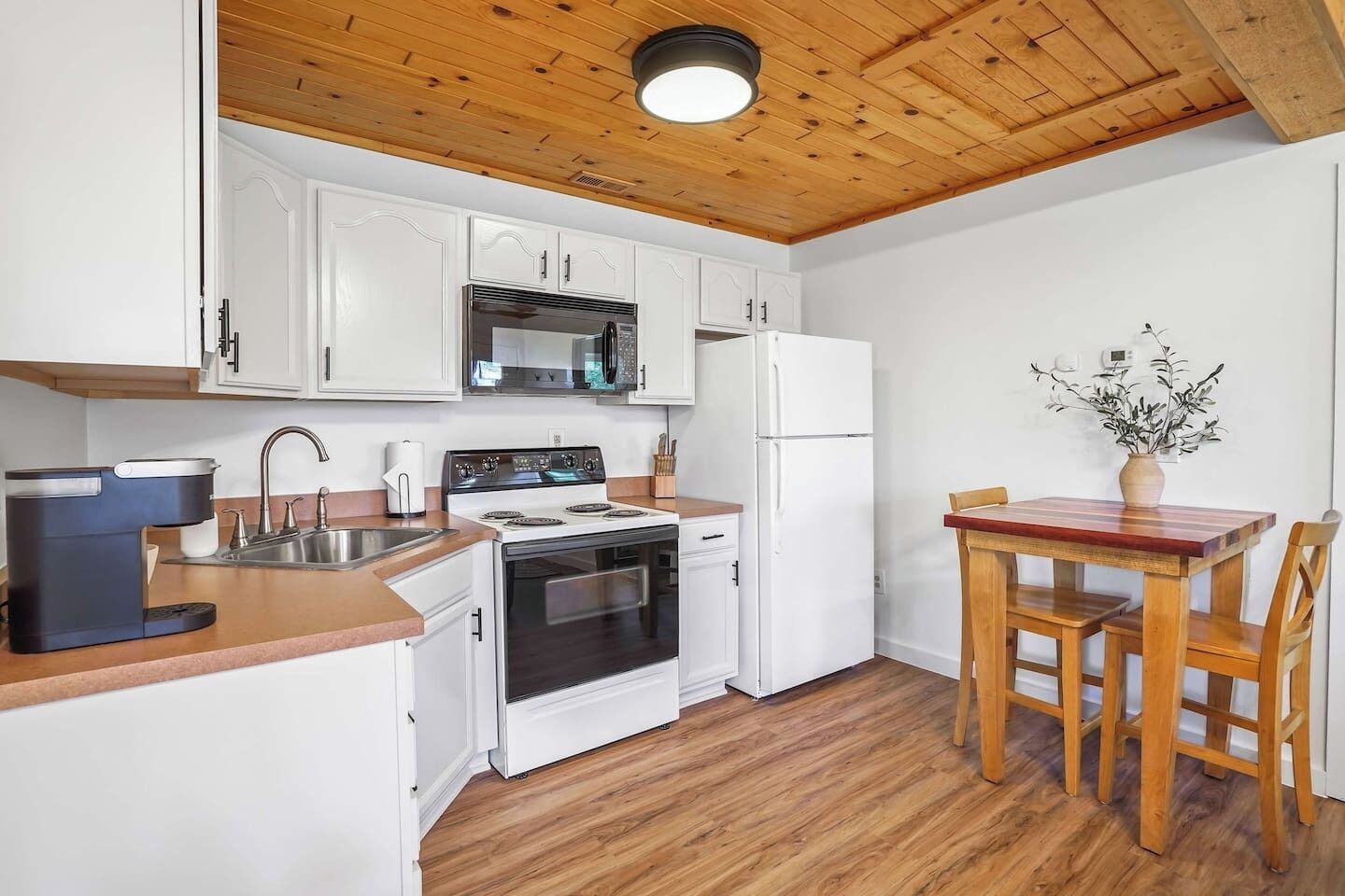 A compact kitchen with white cabinets, a stove, a microwave, and a refrigerator. There's a wooden dining table with chairs and a vase with flowers on the wall.