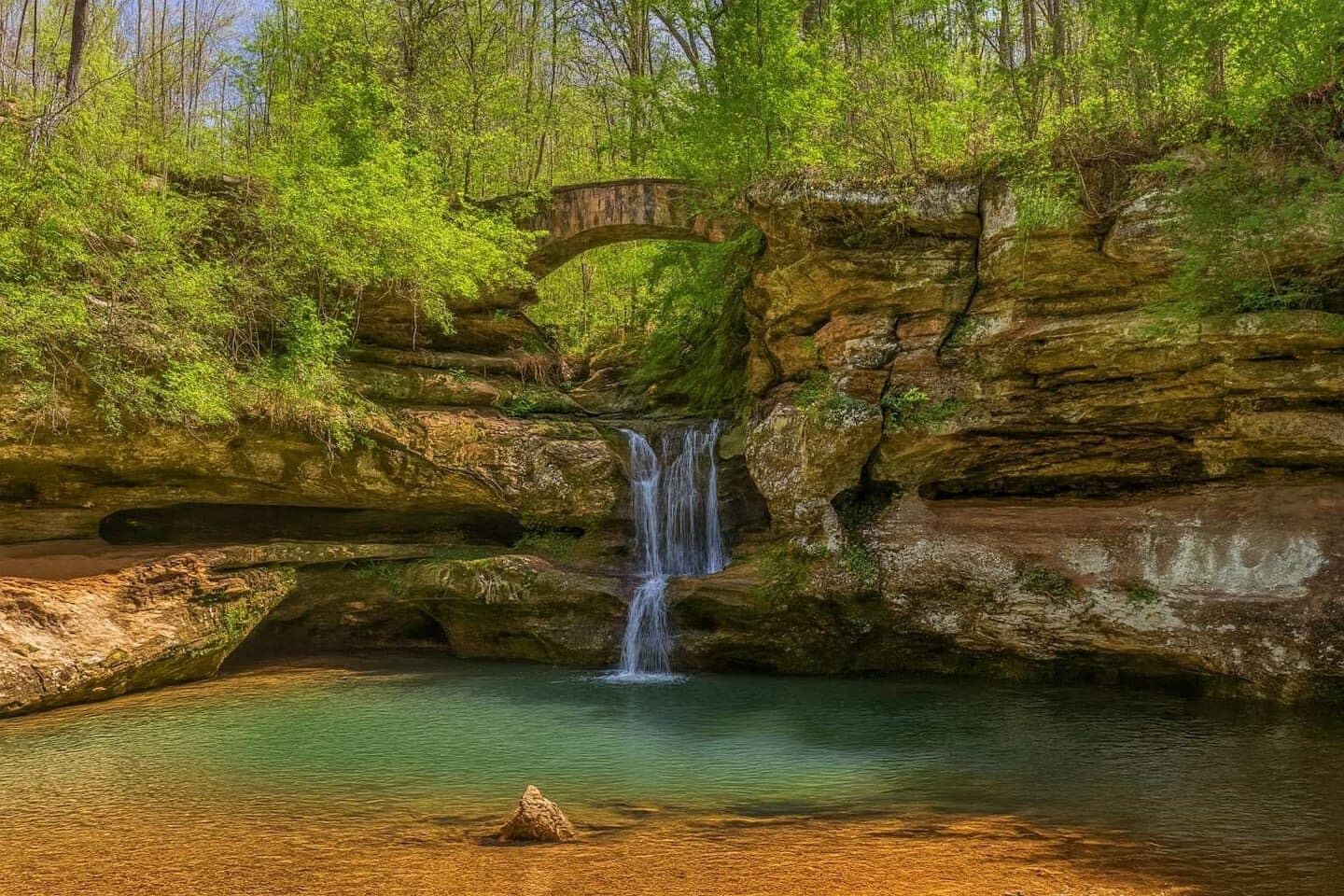 A natural landscape with a waterfall, a small bridge, and a clear pool of water.
