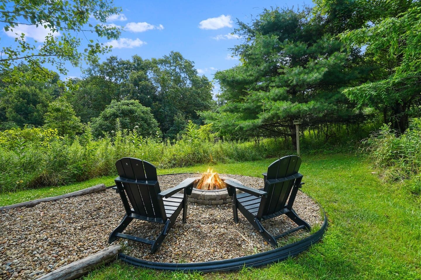 A circular outdoor seating area with two Adirondack chairs around a fire pit, surrounded by gravel and greenery.