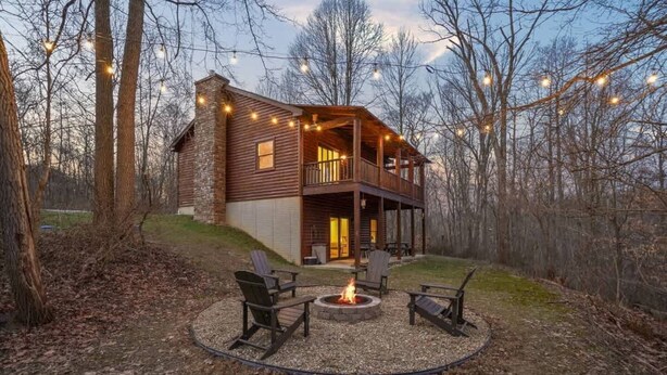 A two-story log cabin with a deck, surrounded by trees and a fire pit with chairs.