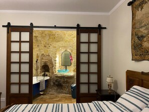 A bedroom with a bed, a nightstand, a lamp, a bathroom with a bathtub, a sink, and a mirror.