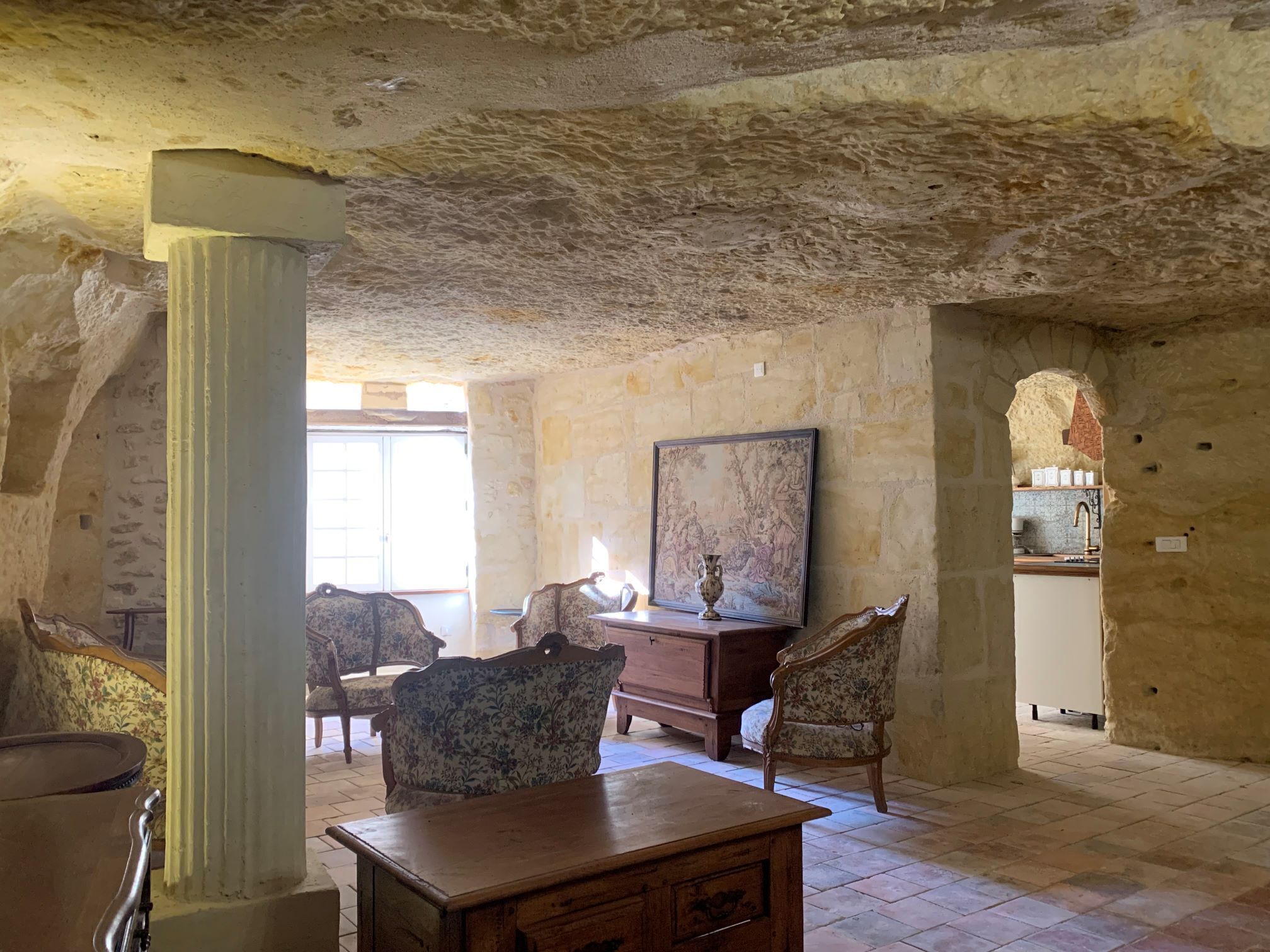 Une pièce aux murs de pierre, avec une table en bois, deux fauteuils rembourrés, un grand tableau encadré et une fenêtre laissant entrer la lumière naturelle.