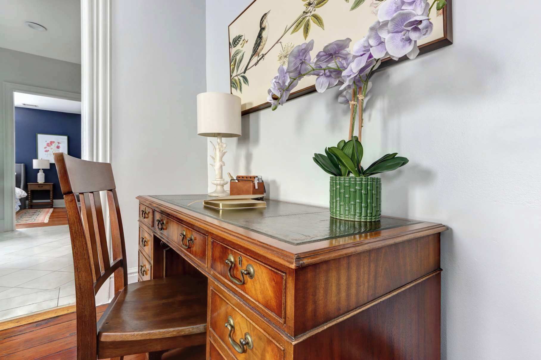 Un bureau en bois avec des tiroirs, une chaise, une lampe et une plante en pot, le tout placé devant un mur orné d’un tableau floral encadré.