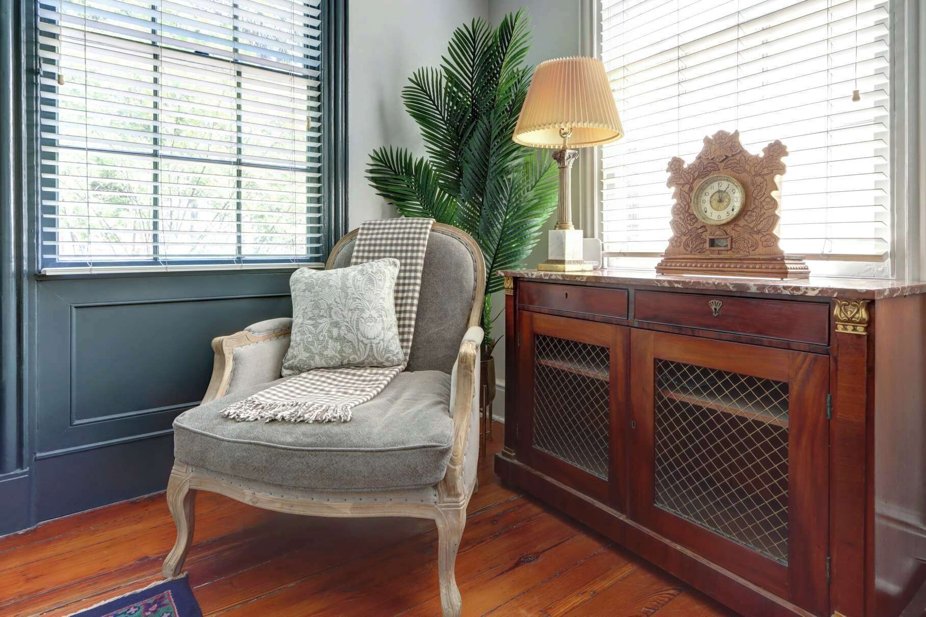 A room with a wooden chair, a lamp, a clock, and a plant.