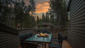 Een houten terras met een glazen tafel en stoelen, omringd door hoge bomen en een huis op de achtergrond.