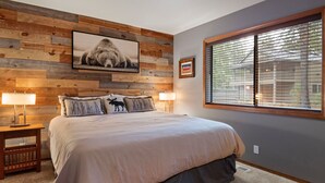 Een slaapkamer met een groot bed, houten wandpanelen, een ingelijst schilderij, twee bedlampen en een raam met jaloezieën.