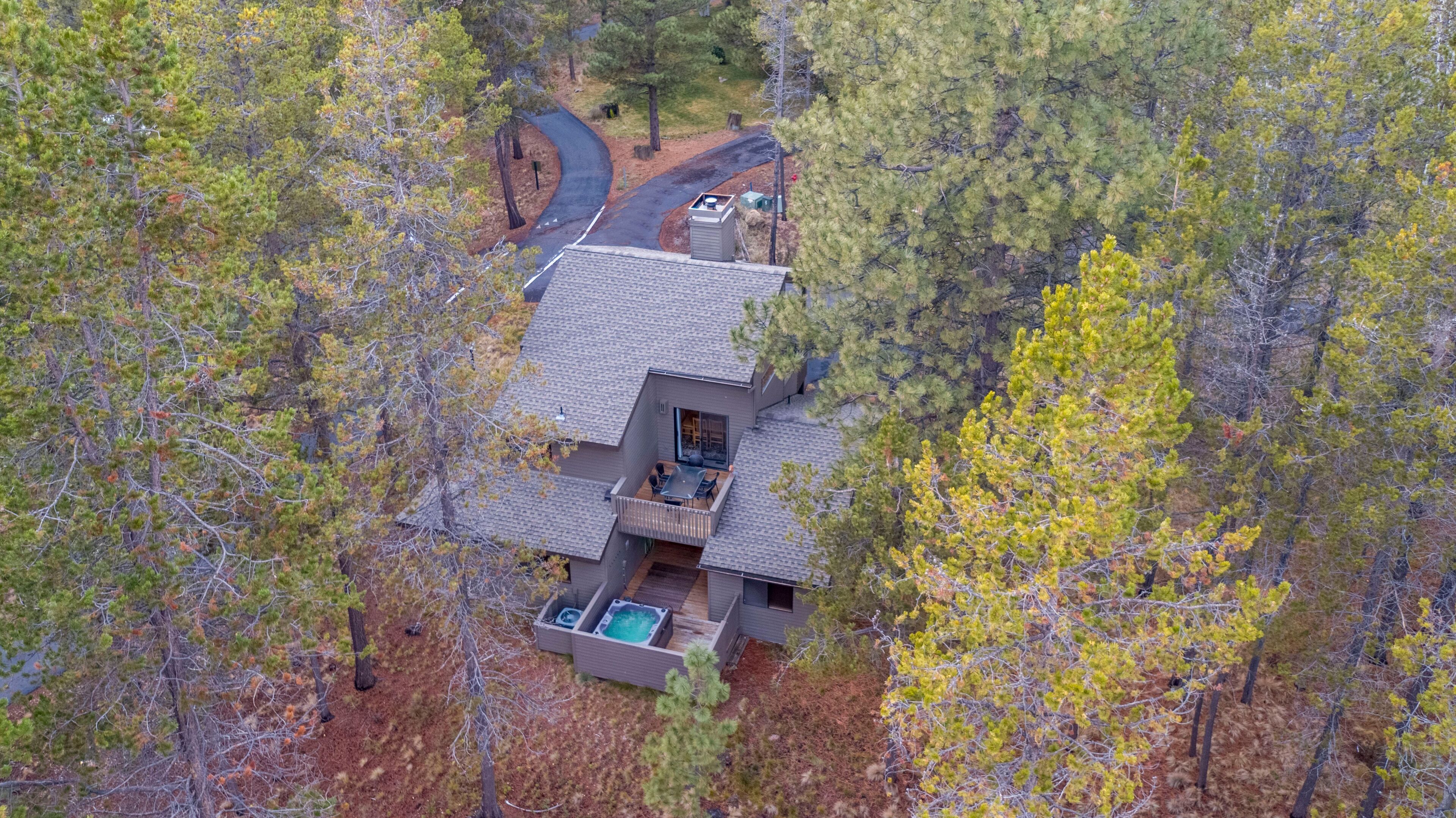 An aerial view of a house surrounded by trees, featuring a large deck with outdoor furniture and a hot tub.