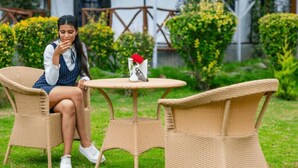 A person sitting at a round table with a vase of flowers, surrounded by wicker chairs in an outdoor setting with greenery and a building in the background. - RishiBhumi Wellness Retreat - Manali (Kullu)