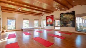 A spacious room with wooden ceilings and large windows, red mats on the floor, and artwork on the walls. - RishiBhumi Wellness Retreat - Manali (Kullu)