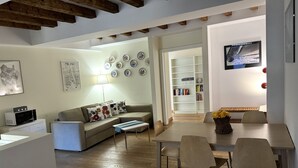 A modern living room with a sofa, coffee table, and dining area with chairs. There is a bookshelf and a TV mounted on the wall. - 1-bed Flat Near The Duomo (Milan)