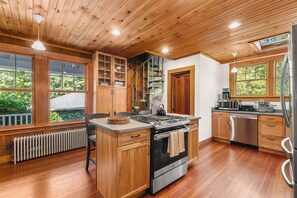 A modern kitchen with wooden cabinetry, a central island, stainless steel appliances, and large windows offering views of greenery. - Tidebend: River Views and Gracious Comfort (Gloucester)