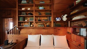 Een knus ingerichte kamer met een houten bed, een boekenplank vol boeken en decoratieve objecten, en een tafeltje met een lamp.