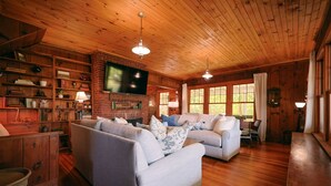 A cozy living room with wooden floors, a fireplace, a flat-screen TV, and a bookshelf. - Tidebend: River Views and Gracious Comfort (Gloucester)