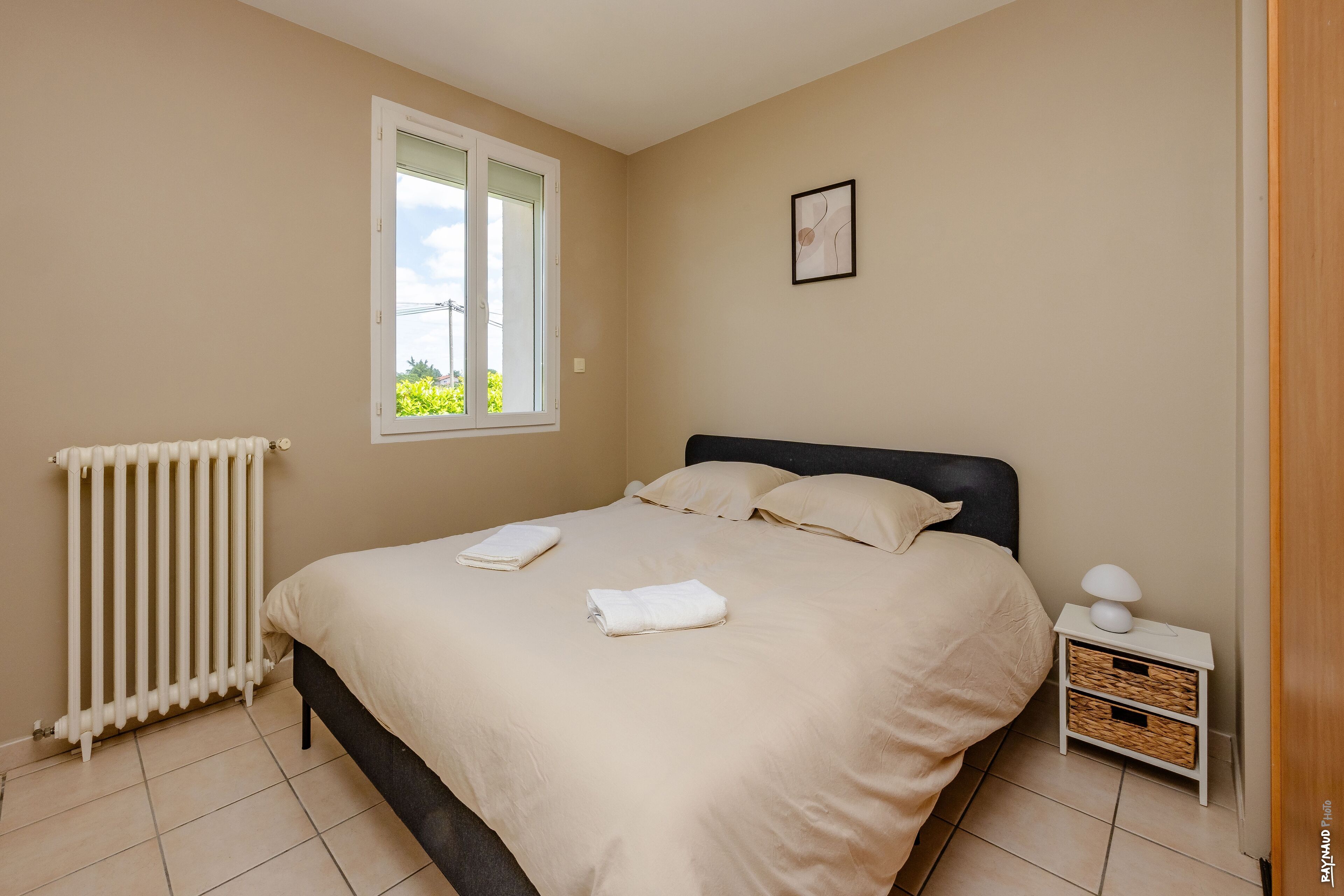 Une chambre à coucher avec un lit, une table de chevet avec une lampe, un radiateur et une fenêtre donnant sur un paysage verdoyant.
