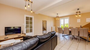 Uma sala de estar moderna com um sofá de couro preto, uma área de jantar com mesa e cadeiras, e uma televisão sobre um móvel de madeira.