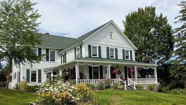 A two-story house with a front porch, surrounded by a well-maintained lawn and flowering plants. - 10 Mi To Gore Mountain: Home In Adirondack Park 5 Bedroom Home (Olmstedville)