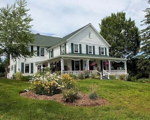 A two-story house with a front porch, surrounded by a well-maintained lawn and flowering plants. - 10 Mi To Gore Mountain: Home In Adirondack Park 5 Bedroom Home (Olmstedville)