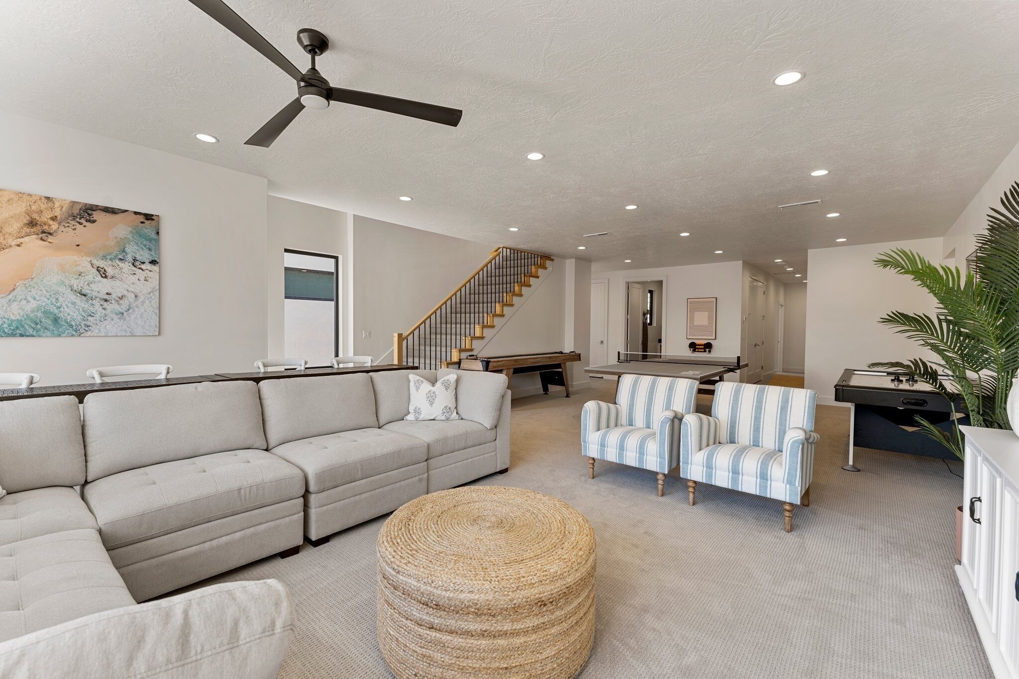 A spacious living room with a sectional sofa, a striped armchair, a woven ottoman, and a ceiling fan.