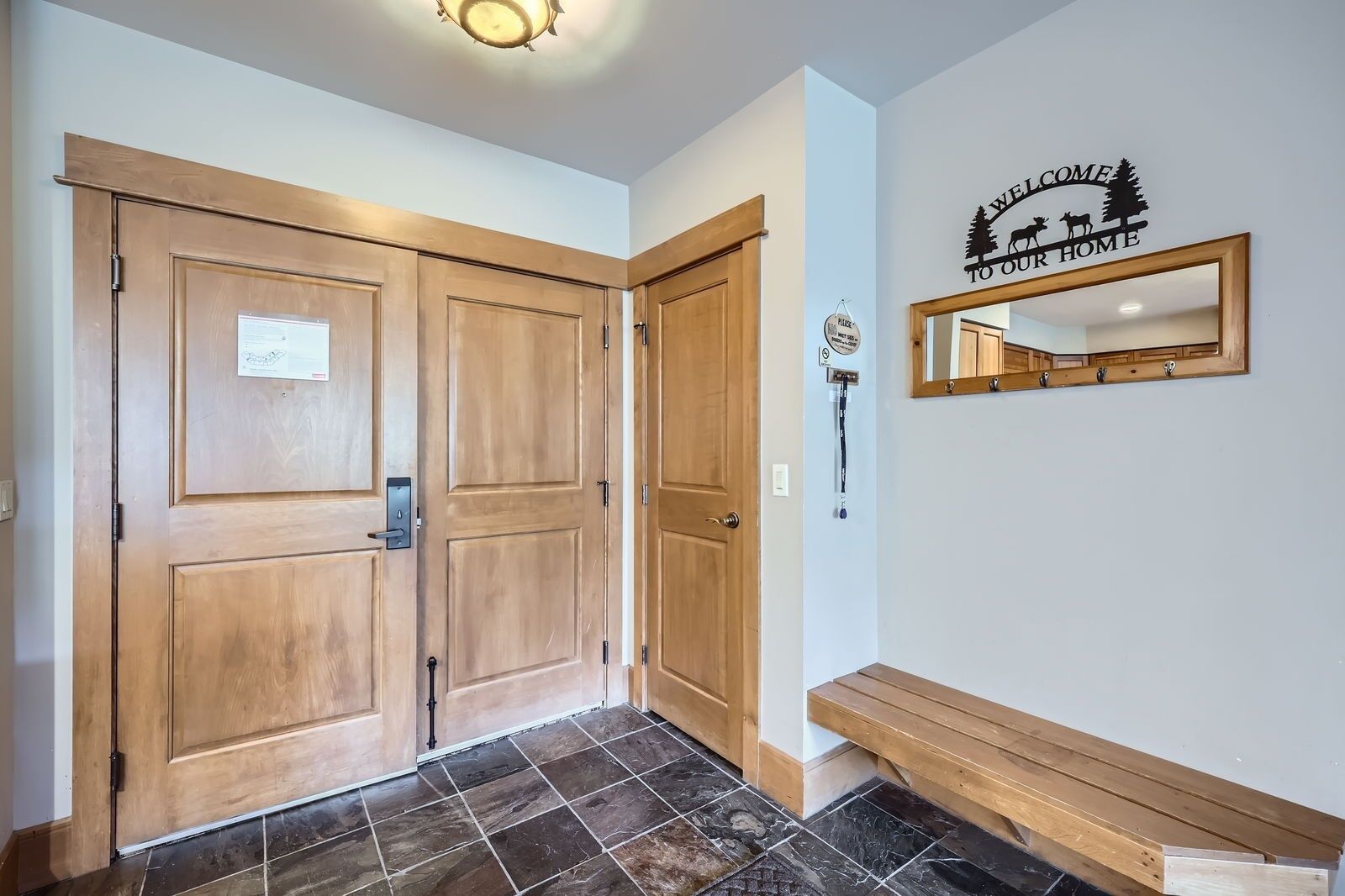 A hallway with two wooden doors, a bench, a welcome sign, and a mirror on the wall.