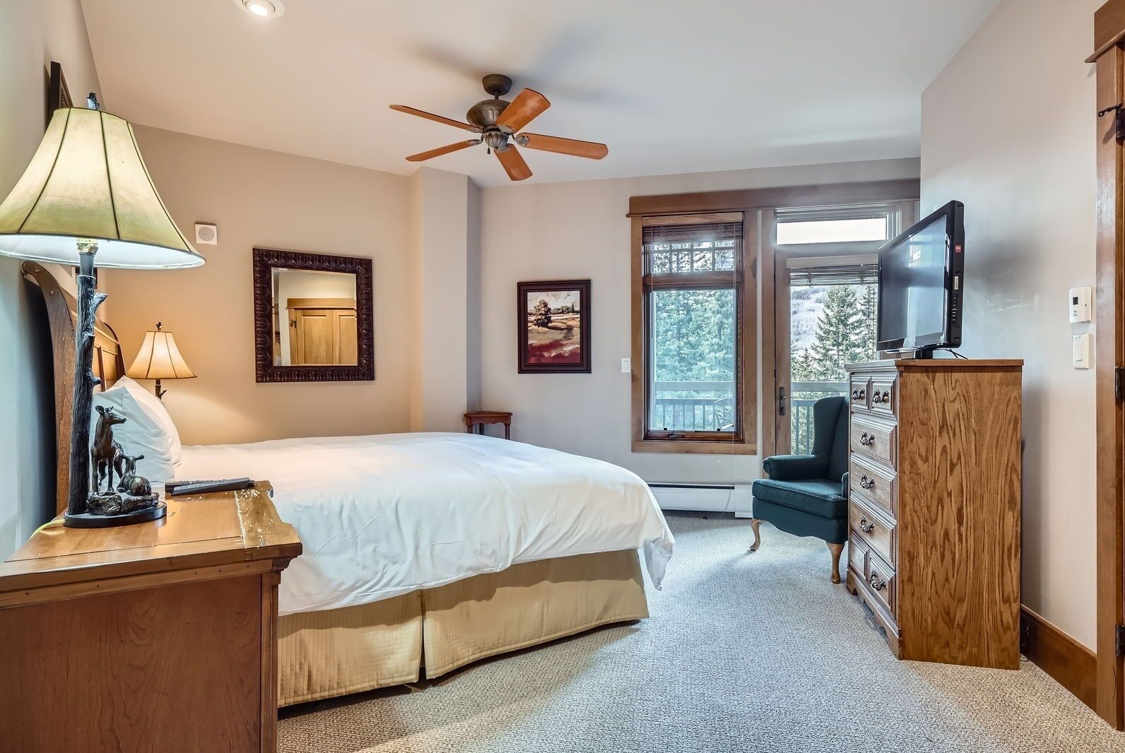 Une chambre à coucher comprenant un lit, un ventilateur de plafond, une commode, un téléviseur et une fenêtre donnant sur des arbres.