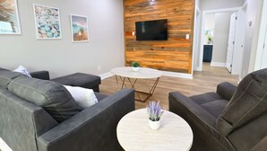 A modern living room with a sofa set, a round coffee table with a plant, and a flat-screen TV mounted on a wooden wall.