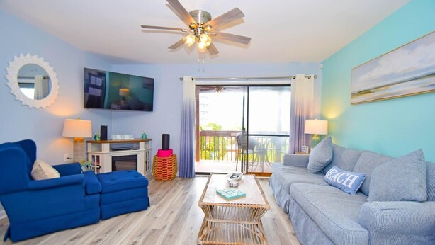 Un salon avec un canapé bleu, un canapé gris, un ventilateur de plafond, un téléviseur et un balcon avec vue.