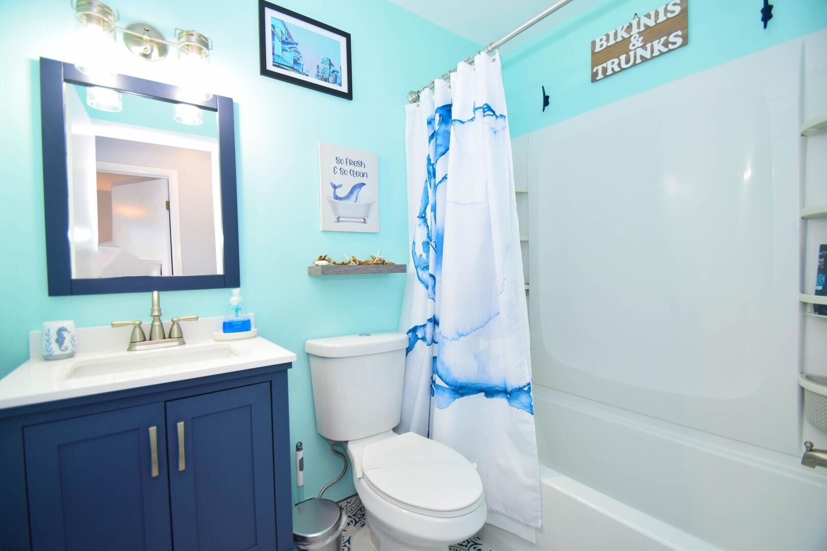 Un baño con tocador azul, inodoro blanco y cortina de ducha con diseño azul y blanco. Se observa un letrero que dice "Bikinis & Trunks".