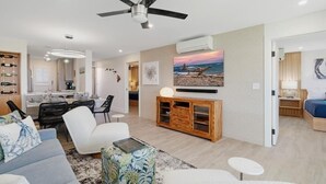 A modern living room with a ceiling fan, a dining area with a table and chairs, a kitchen with a coffee machine, and a bedroom with a bed and a nightstand. - Menehune Shores, #119 2 Bedroom Condo (Kihei)