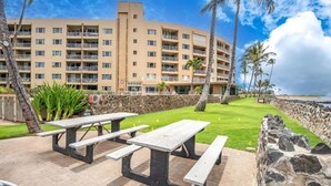 A multi-story hotel with balconies, a lawn area with picnic tables, and a view of the beach. - Menehune Shores, #119 2 Bedroom Condo (Kihei)