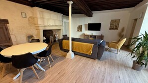 A spacious living area with a round dining table, black chairs, a grey sofa with a yellow throw, and a large potted plant.