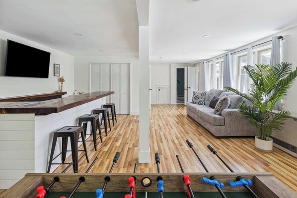Un salon moderne avec un baby-foot, un canapé gris et une grande fenêtre avec des rideaux.