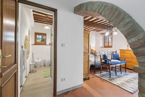 Une pièce avec une porte en bois, une arche en pierre, un lit avec une tête de lit en bois, une commode et une chaise avec un coussin bleu.