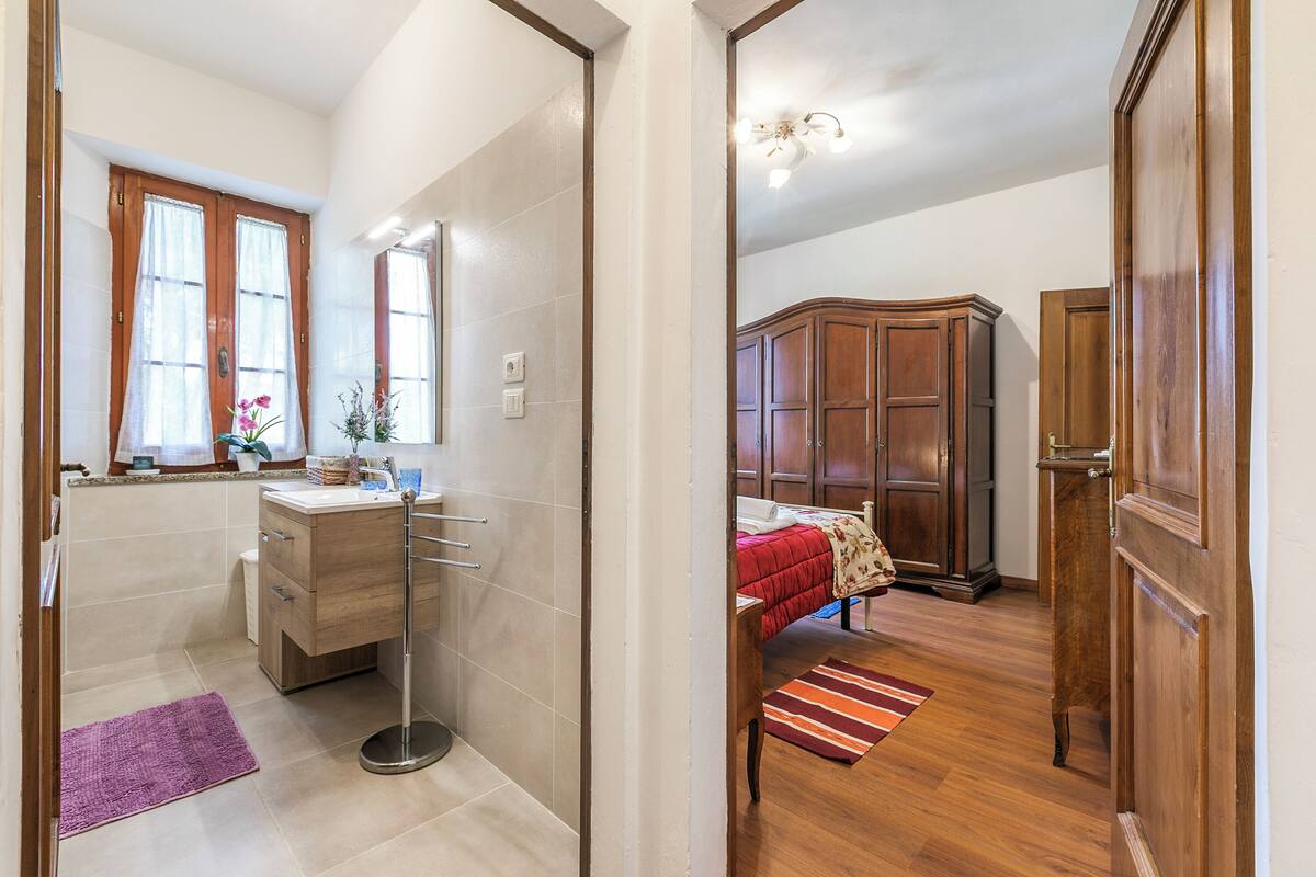 A bedroom with a wooden wardrobe, a bed with a red blanket, and a bathroom with a sink and mirror.