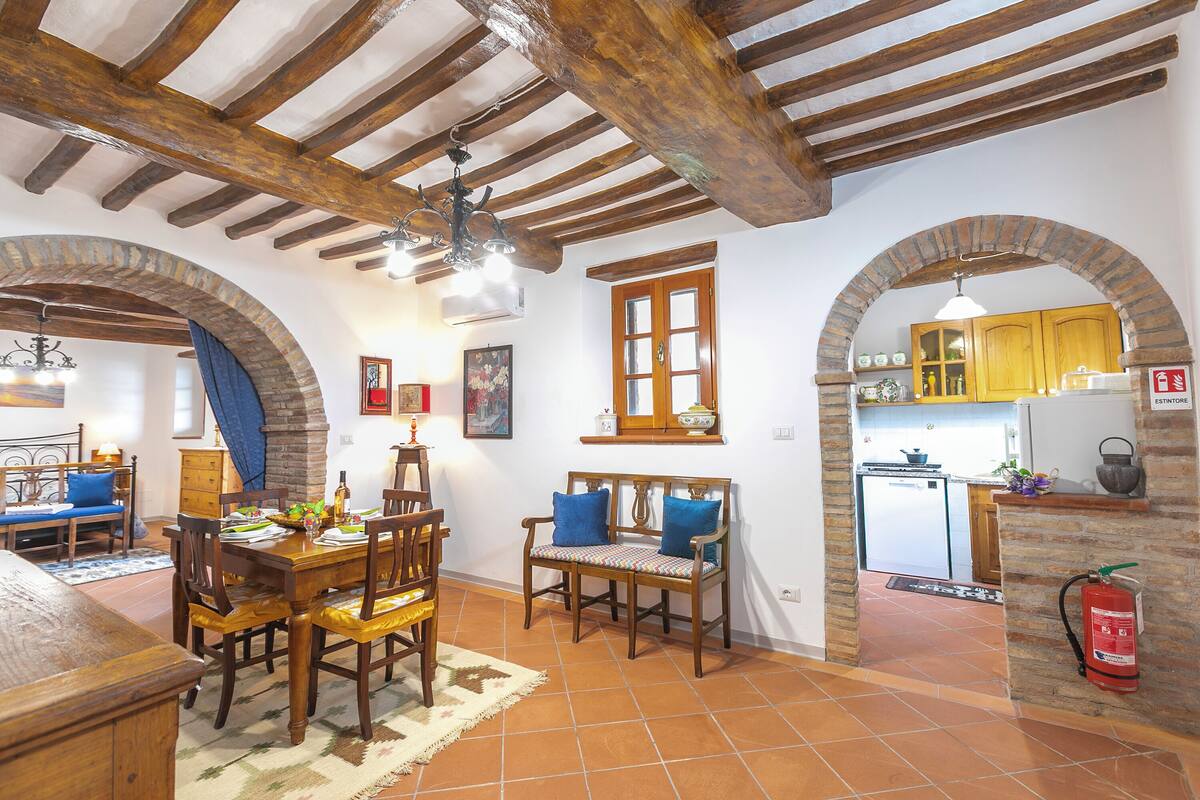 A dining area with a wooden table and chairs, a bench, and a kitchen visible through an archway.