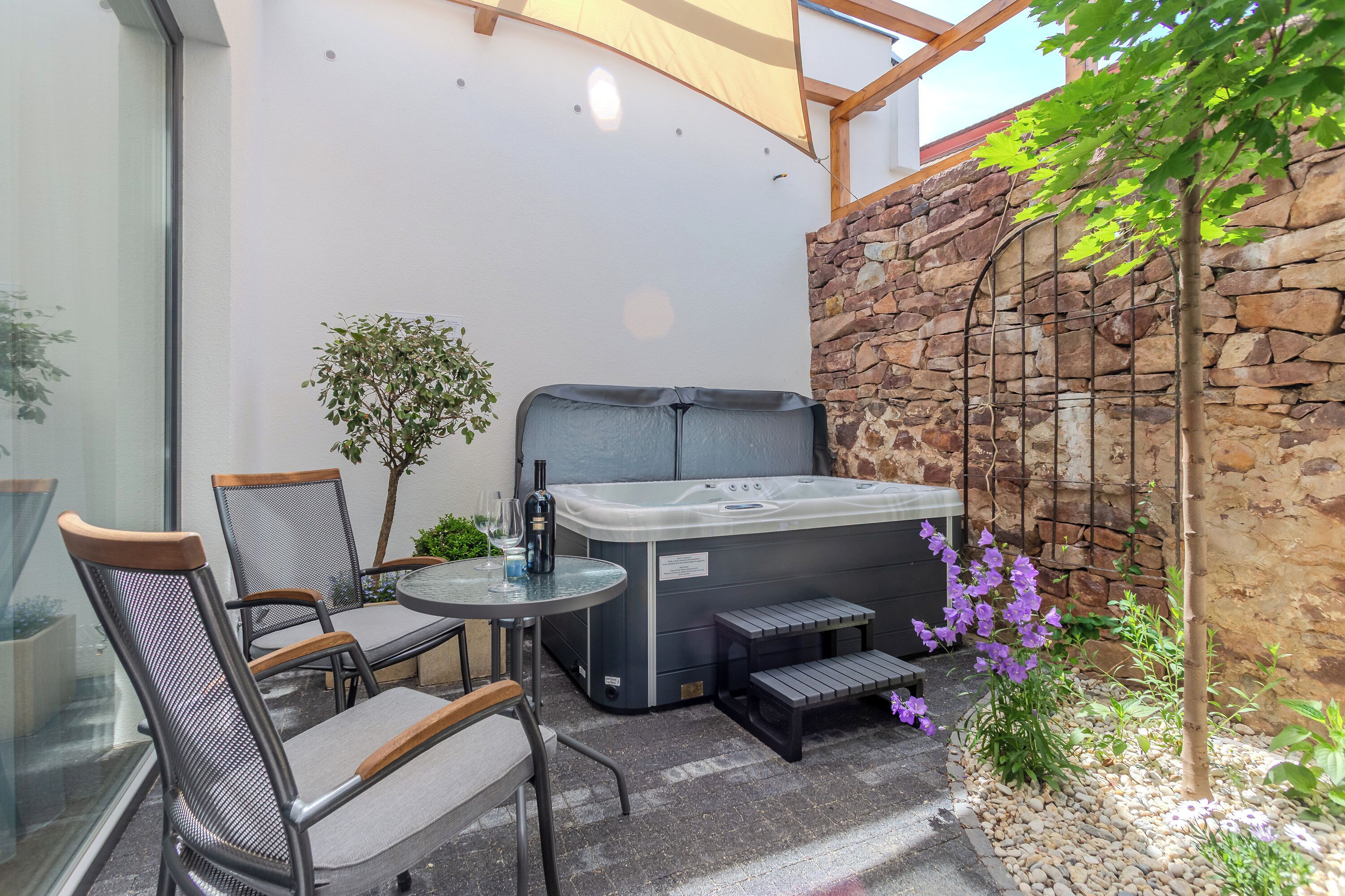 A patio with a hot tub, chairs, a small table, and a stone wall.